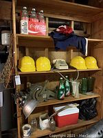 Wooden shelves containing five yellow hard hats, two vintage rotary telephones in cream, vintage Coca-Cola and Canada Dry 1-liter glass bottles, green glass vases, box sealer, metal lamp, and red cooler.