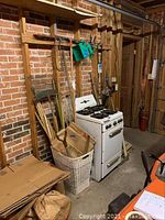 Wide shot showing multiple garden tools including rakes, a stove, and a basket of lawn bags leaning against a brick wall in a basement area.