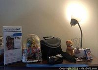 Various items on a table including a WonderArt latch hook kit, a snow globe with Christmas decor, camcorder carrying case, small teddy bear, photo holders, small decorative box, and lamp