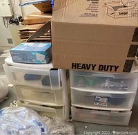 Two plastic storage containers placed side-by-side, filled with multiple items inside translucent drawers; a cardboard box and other packaging materials are on top and nearby.