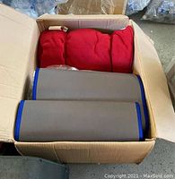 Boxes contain folded gray Korean sleeping mat with blue edge trim and a white Korean character. Red blankets rolled or folded beside the mat inside a cardboard box.