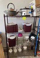Wide view of metal shelving rack holding the wok with glass lid, Hamilton Beach slow cooker, multiple maroon and white plastic storage containers, beverage dispenser, clear glass jars, and green bag on bottom shelf.