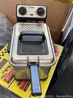 Close-up of T-Fal deep fryer showing control panel with dials and black handle on lid. Stainless steel body visible.