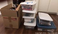 Six clear plastic storage bins stacked, containing various craft and office supplies next to a large cardboard box.