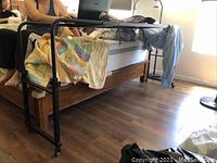 Photo showing the full over-the-bed desk frame partially positioned over a wooden bed with bedding draped around it. The structure is black metal with caster wheels on each leg for mobility.