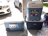 Three ice chests grouped in a garage: two with wheels and one without. Heights and colors visible.