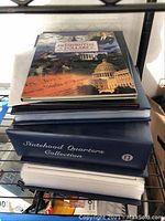 Stack of three collectors’ albums: Presidential Dollars on top, Statehood Quarters box beneath, and Historic Walking Liberty folder at bottom