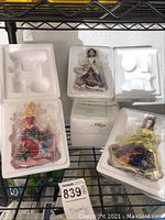 Seven Princess Belle figurines in white styrofoam packaging on a metal shelf, sealed in plastic, some certificates visible below.