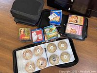 Close-up of two black zippered binders with CDs/DVDs inside, several loose DVDs and Blu-rays on floor including titles such as Big Hero 6 and Epic