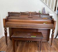 Full front view of wooden upright piano with matching bench underneath. Piano has turned legs and carved music stand.