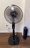 Amazon Basics black pedestal fan with metal cage and visible control buttons on pedestal base next to a smaller black tower heater.