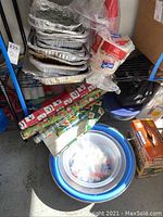 Stacked aluminum trays with red disposable cups and paper plates and napkins in packaging, gift wrap rolls and plastic bowls on floor