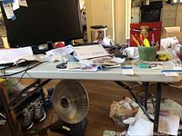 Folding table with white top supported by black metal legs, cluttered with assorted office and school supplies, cables, papers and a small fan on the floor beneath the table.