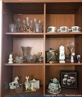 Full view of three shelves in wooden cabinet with multiple glassware, ceramic tea cups, candle holders, salt and pepper shakers, and decorative figurines