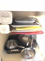 Kitchen cabinet with stacked baking trays, cooling racks, cutting boards in red and yellow, two rectangular food storage containers, and metal pots and pans including one with a glass lid.
