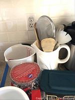 Kitchen items on a counter including a measuring cup, white pitcher with wooden and plastic utensils, and a dark green container lid.