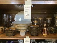 Full shelf showing the Noritake plates and bowls stacked in front, crystal glassware with gold rims behind, and decorative bell beside them