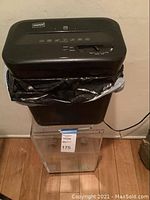 Photo showing black Staples paper shredder atop clear acrylic storage bin with black liner inside, placed on wooden floor.
