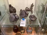 Photo showing six small dark brown ceramic decorative teapots and sculptures shaped like animals and abstract figures inside a glass display.