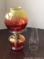 Oil lamp with combined red and amber colors, spherical base and globe, and an unused wick. Separate clear glass chimney placed beside it.