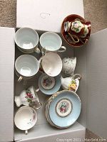Top-down photo showing a box containing a variety of china cups and matching saucers with floral patterns and gold accents.
