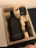 Opened box displaying black Saje AromaOm ultrasonic diffuser, power adapter, and two bottles of oil in molded cardboard packaging.