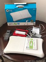 Red Wii console, white Wii Fit balance board with Wii Fit Plus game and white Wii Remote on top, sensor bar, power adapter and charging stand with cables shown on table.