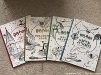 Four Harry Potter collectible colouring books laid out in a row on a fabric surface showing front covers with titles and illustrations on light-colored backgrounds with red, gold, green, and blue accents.