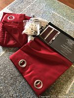 Photo showing two sets of red curtains, one unopened in packaging and one folded, along with black and white shower curtain and hooks on floor.