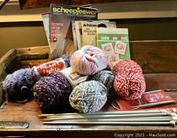 Photo showing six balls of yarn in different colors and textures with knitting pattern books in the background.