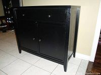 Black cabinet angled view showing two doors, one drawer, and cabinet legs on tiled floor