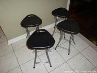 Four black plastic seated folding stools with triple tubular silver metal legs on a tiled floor against a wall.