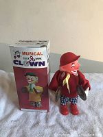 Photo of the vintage mechanical wind-up musical cymbal clown next to its original box showing the clown in red cap and jacket with cymbals.