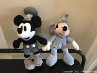Photo showing Steamboat Mickey and Elephant Mickey plush dolls sitting side by side on a dark surface.