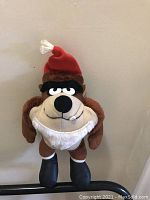 Brown and beige plush Taz character wearing a red Santa hat standing against a beige wall.