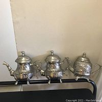 Photo of three silver-toned metal teapot wall decors with intricate details and mounting hooks, side by side against a plain background.