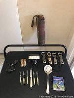 Photo of all items in lot displayed on a black surface with handle behind showing umbrella and grouped utensils and openers on black chair.