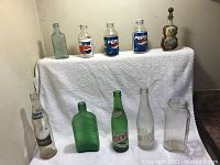 Nine vintage beverage bottles arranged on and in front of a white towel. Includes Pepsi, Fanta, Elders, a violin-shaped liquor bottle, and others.