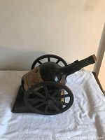 Side view of the large dark glass Courvoisier cognac bottle mounted on a dark cannon-style wooden carriage, on a white cloth background.