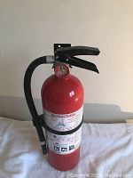Red fire extinguisher against plain wall, showing front with labels, hose, handle and pressure gauge.