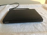 Photo showing the slim black Sony DVD player placed on a light cloth surface. Front view captures the logo and front panel buttons.