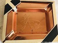 Top view of a copper-colored rectangular metal ashtray engraved with crossed curling brooms, a curling stone, and target rings, with slots on the corners for resting cigarettes.