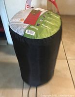 Black cylindrical carrying pouch with red strap handle holding Coleman Classen sleeping bag. Package top label visible with product information.