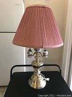 Brass table lamp with pink pleated fabric shade and decorative ceramic accents on lamp body, placed on a black surface.