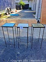 Front view of all three garden plant stands together outside on pavement showing weathered condition and black metal base with colorful ceramic tops.