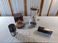 Photo showing four kitchen items on table: Cuisinart spice grinder, KitchenAid blender, glass dome cake stand, and sealed boxed item