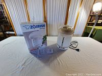 White Zojirushi electric air pot CW-PZC22 and its original box placed on a white covered surface with wood-frame background.
