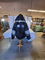 Front view of black Raiders hooded jacket on stand, showing Raiders logo patch on chest and silver sleeve stripes.