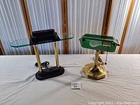Two vintage desk lamps on white cloth. Left lamp is black and brass with glass shelf, right lamp is green glass shade banker lamp with brass base.