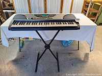 Full front view of Yamaha PSR-240 keyboard resting on a white covered table with an adjustable black X-style metal stand supporting it.